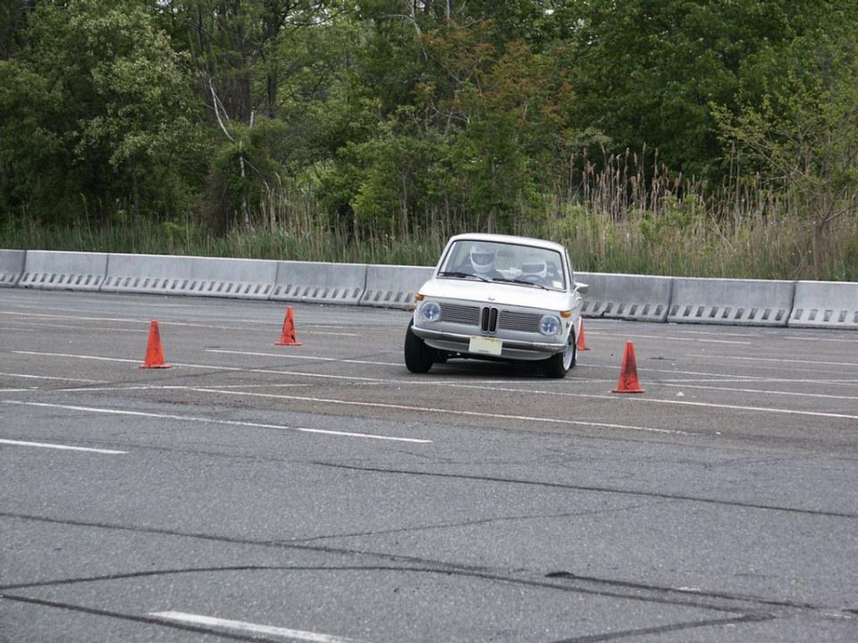 White BMW 2002 with driver wearing helmet navigates an autocross course with orange cones