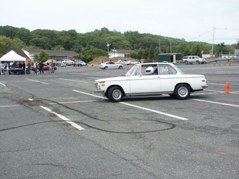 2001 Mercury Grand Marquis - A white coupe with a driver in a helmet participates in an autocross event, with other vehicles and tents visible in the background.
