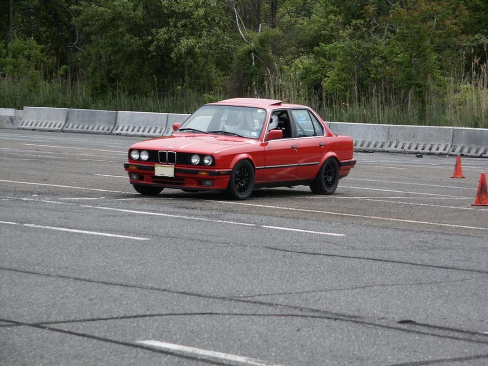Red sedan with black wheels on a track, driver visible inside. Orange cones are on the side