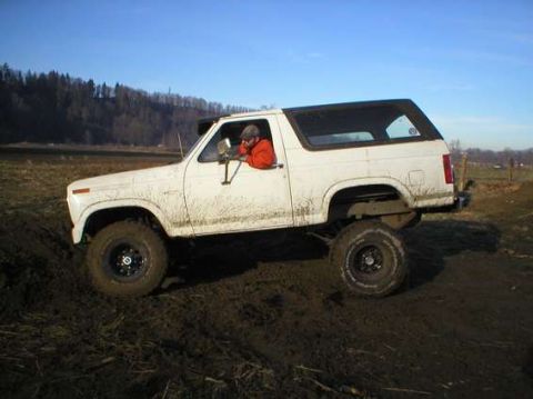 1979 Ford Bronco - A white SUV navigates a muddy terrain under a clear blue sky with trees in the background.