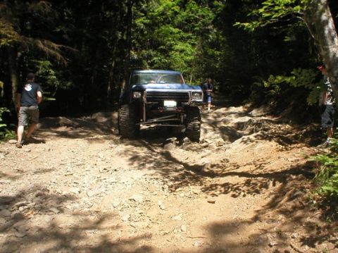 1979 Ford Bronco - A modified blue SUV navigates a challenging, rocky off-road path surrounded by dense forest.