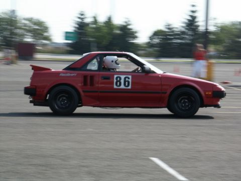 2001 Mercury Grand Marquis - A red sports car, identified by the number 86 on its door and a driver in a white helmet, is captured in a side profile during a motorsport…