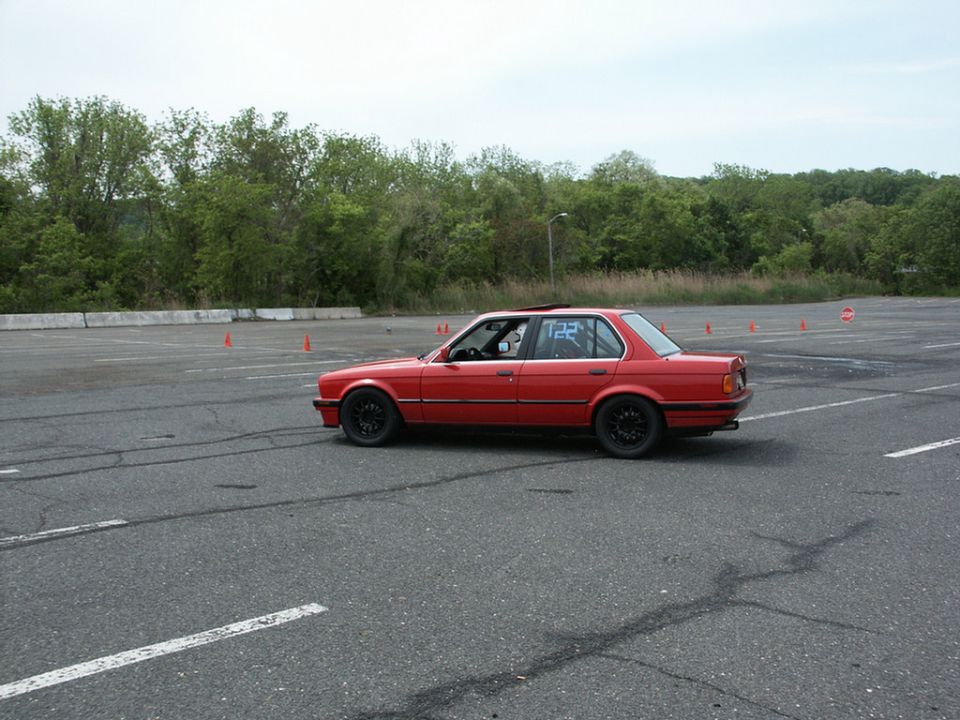 Red sedan with black wheels and racing number 122 on the rear window, parked on asphalt with orange cones in the background