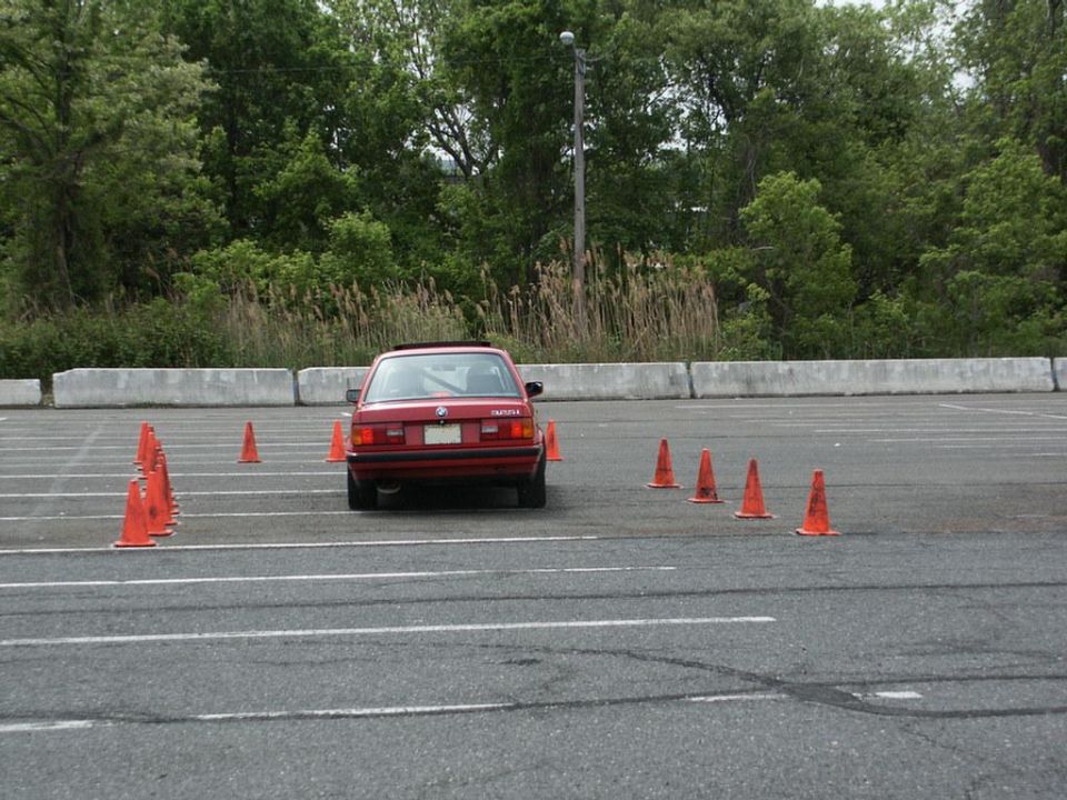 Red BMW E30 sedan navigating an autocross course marked by orange cones