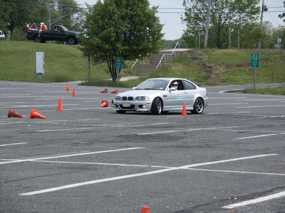 Silver BMW M3 coupe with number 168 on door, navigating a cone course. Dark pickup truck with passengers is visible on a grassy hill in the background