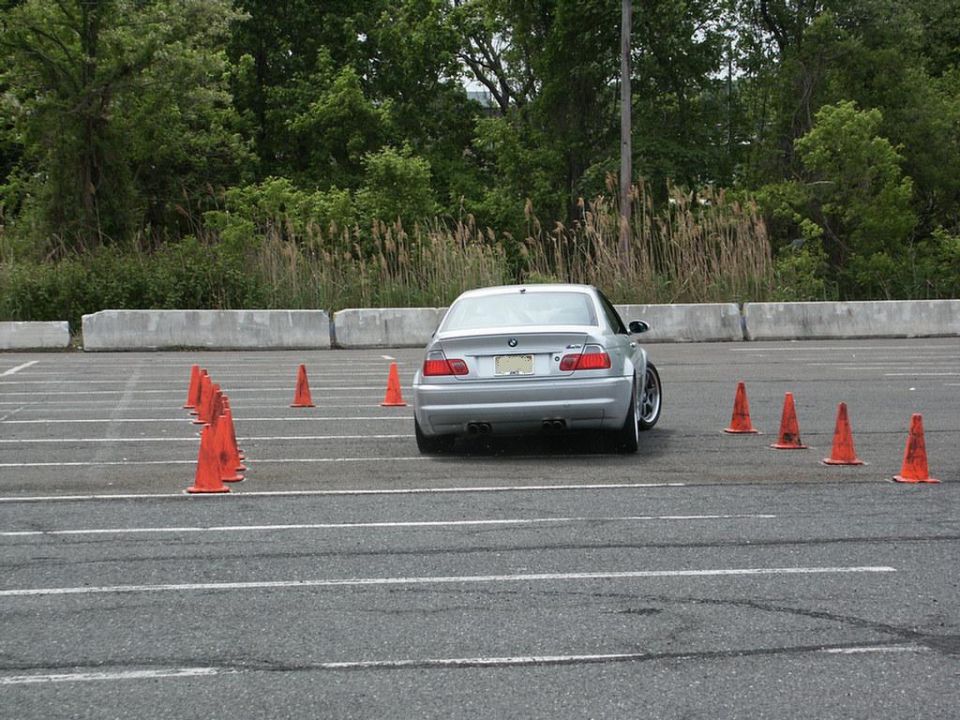 Silver BMW M3 performing an autocross maneuver in a parking lot, with orange cones marking the course
