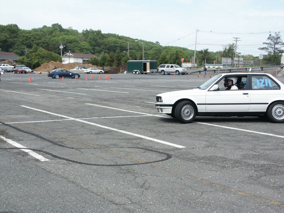 White BMW E30 coupe doing autocross, driver wearing helmet. Other cars and cones visible in background