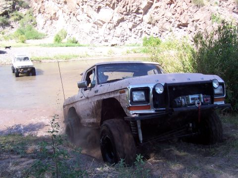 1981 Ford F-150 - Two trucks navigate a shallow riverbed with rocky cliffs in the background.