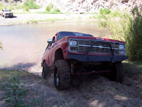 1981 Ford F-150 - A red pickup truck with large tires navigates a shallow river crossing, with another vehicle visible on the bank.