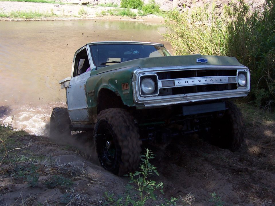Green and white Chevrolet pickup with large tires driving through mud and water. Grille reads CHEVROLET