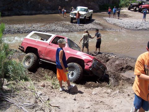 1981 Ford F-150 - A red SUV is stuck in the mud while people attempt to help it out near a river, with other vehicles visible.