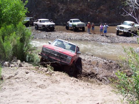1981 Ford F-150 - Several off-road vehicles navigate a river crossing, with one truck deeply mired in mud.