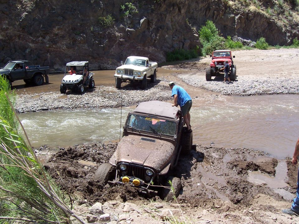 Muddy Jeep stuck in a riverbed, people attempting to push it out. Other vehicles are nearby