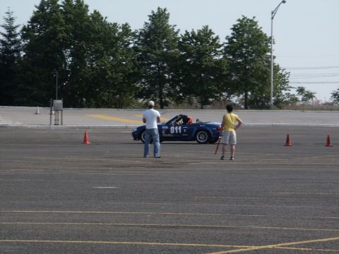 2001 Mercury Grand Marquis - A blue convertible sports car is parked on a lot, with two people observing it. Orange cones are visible around the area.