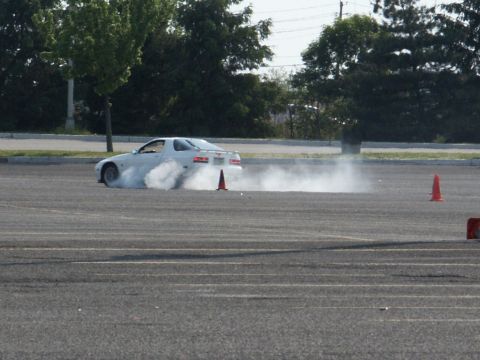 2001 Mercury Grand Marquis - A white sports car performs a drift maneuver, creating a cloud of smoke on an asphalt course marked with orange cones.