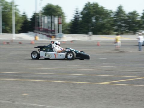 2001 Mercury Grand Marquis - A driver in a helmet pilots a small, open-wheel race car on a paved track with cones and spectators in the background.