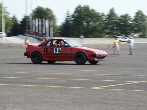 2001 Mercury Grand Marquis - A red sports car with racing decals drives on a paved track with cones and spectators in the background.