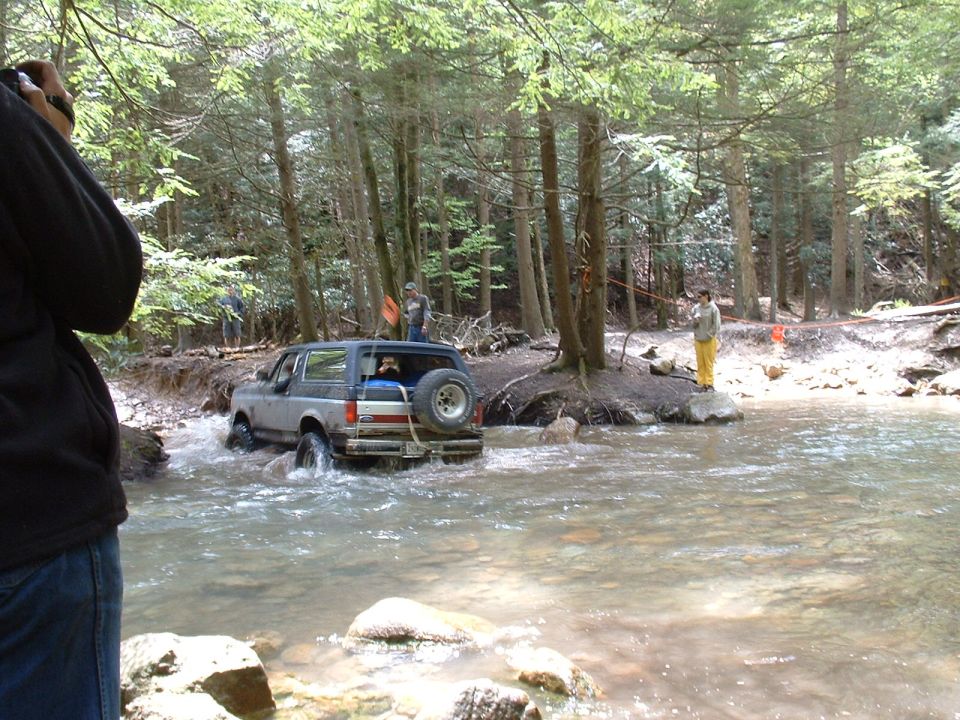 Ford Bronco driving through a stream with trees and people on the bank