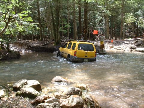 1991 Ford Bronco - A yellow SUV navigates a rocky riverbed surrounded by trees.