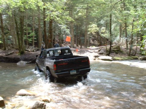 1991 Ford Bronco - Dodge Ram pickup truck navigates a stream in a wooded area.