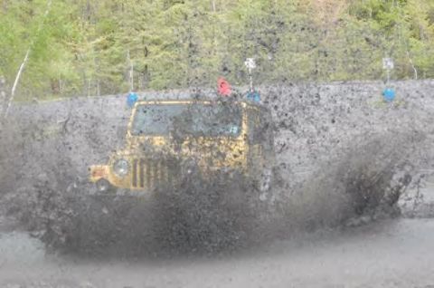1991 Ford Bronco - A yellow Jeep navigates a muddy obstacle, sending mud flying in all directions.