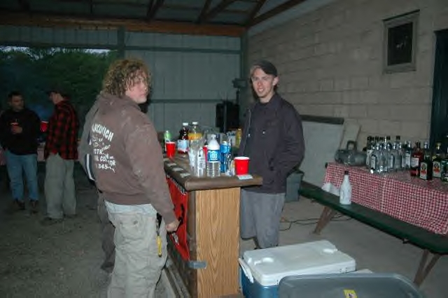 People stand around a bar with drinks and bottles on a table behind it