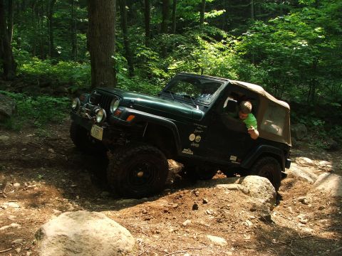 1996 Ford Bronco - A driver navigates a challenging rocky path in a Jeep Wrangler through a dense forest.