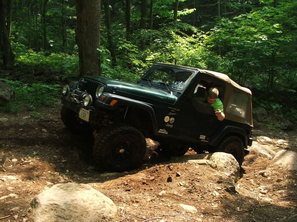 Green Jeep Wrangler driving over rocks on a wooded trail. Driver gives a thumbs up