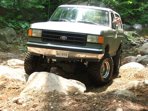 1996 Ford Bronco - A modified Ford Bronco is shown off-roading on a rocky trail with a stream in the background.