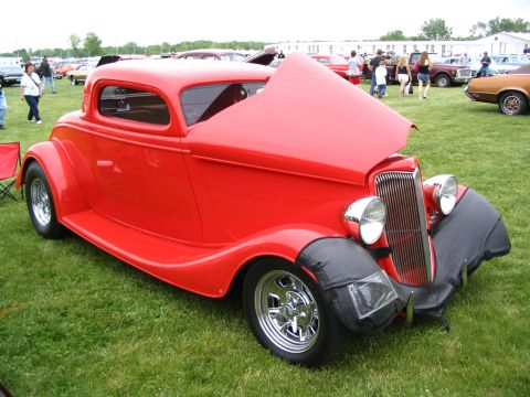 1993 Ford Taurus - A bright red hot rod coupe is displayed with its hood open at an outdoor car show event.