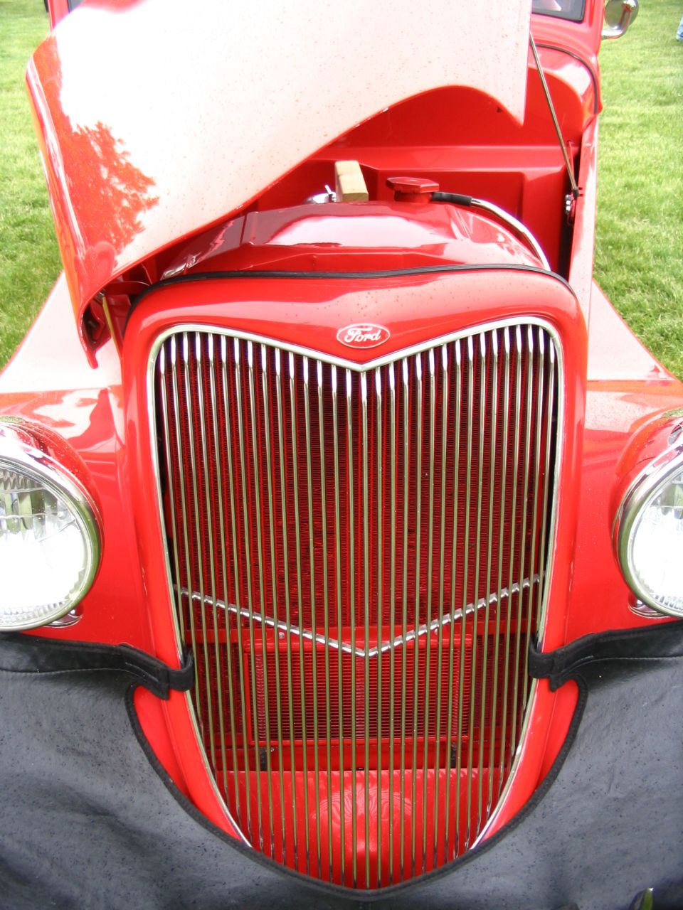Red Ford truck grille with vertical chrome bars and a Ford badge. Hood is open, revealing the engine bay. Headlights are visible on either side