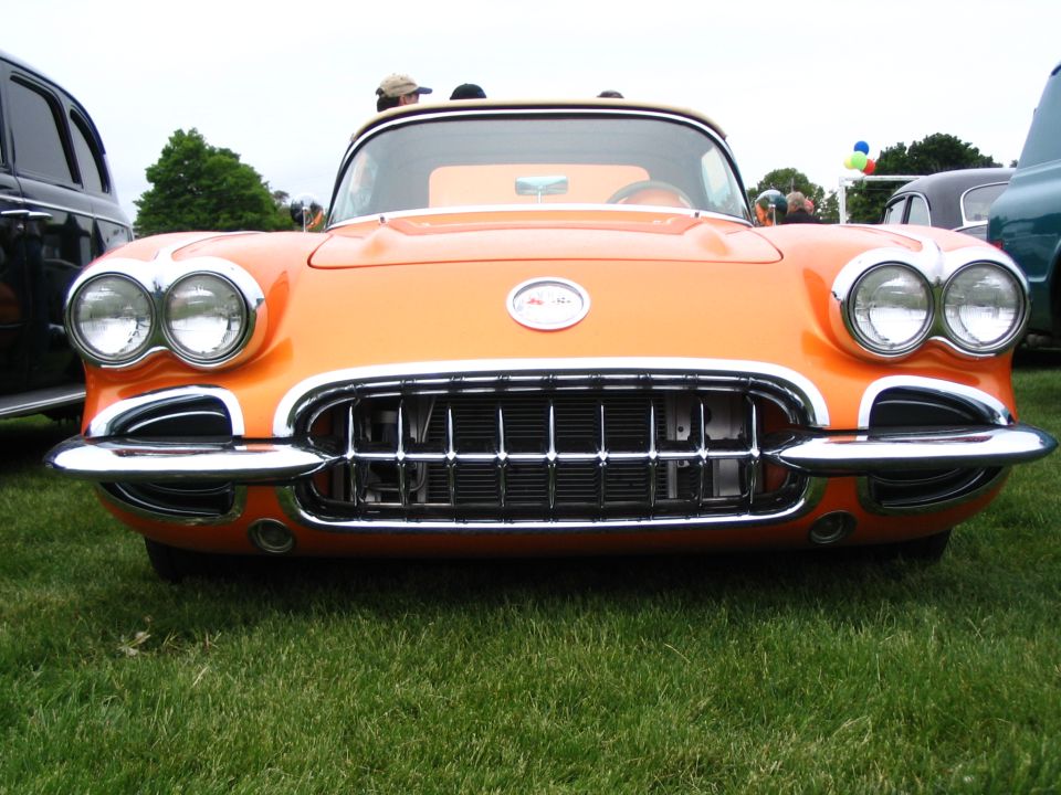 Orange 1958 Corvette front end with chrome grille and headlights. Other cars visible in background