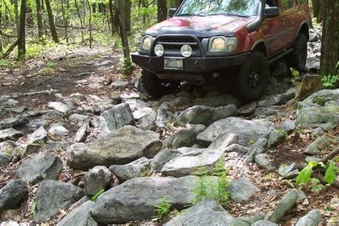 1991 Ford Bronco - Off-roading SUV tackles a challenging, boulder-strewn path in a wooded area.