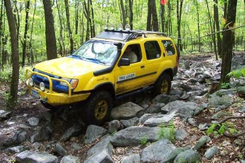 1991 Ford Bronco - A yellow SUV, modified for off-roading with a snorkel and roof rack, tackles a challenging rocky path through a wooded area.