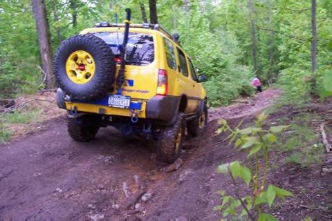 1991 Ford Bronco - A yellow SUV navigates a muddy trail in a wooded area, with another vehicle seen further up the path.