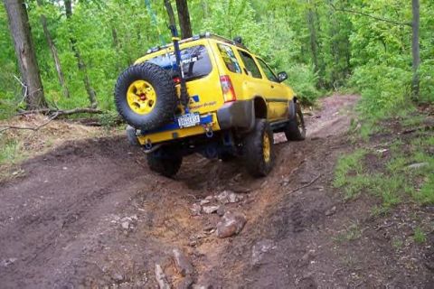 1991 Ford Bronco - Yellow SUV navigates a challenging muddy trail in a wooded area.