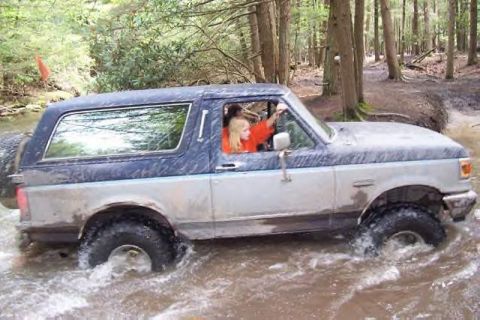 1991 Ford Bronco - A two-tone blue and silver Ford Bronco navigates through a muddy stream in a wooded area.