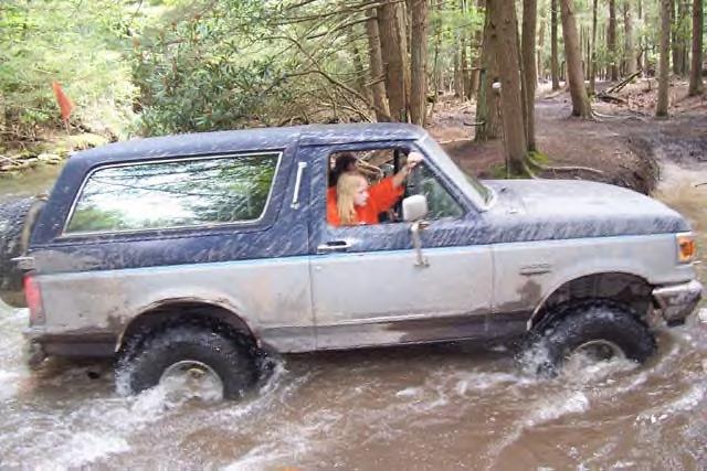 Two-tone blue and silver Ford Bronco driving through shallow water in a forest