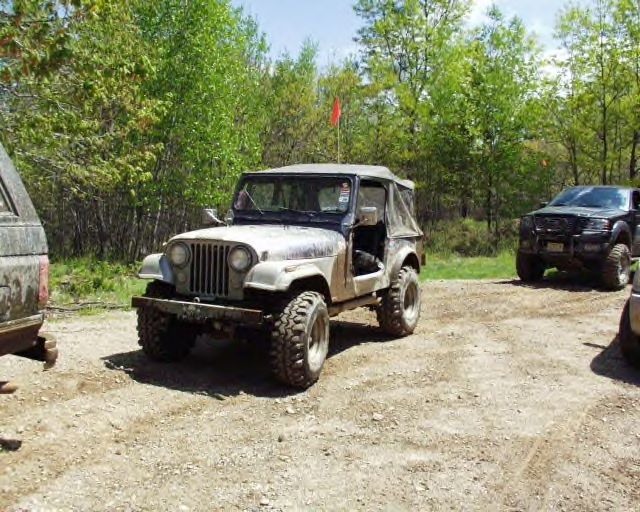 Jeep CJ-5 parked on a dirt road with trees in background. Another SUV is to the right