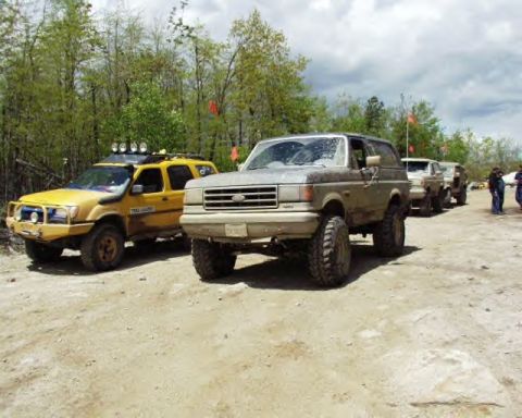 1991 Ford Bronco - A line of off-road vehicles, including a gray Ford Bronco, are parked on a dirt path with trees in the background.