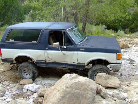 1987 Ford Bronco - Ford Bronco shown off-roading on a rocky path, with visible damage on the driver's side.