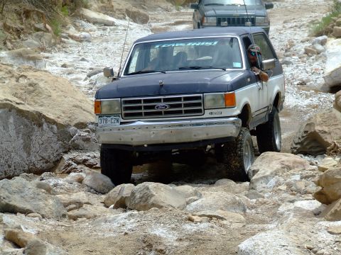 1987 Ford Bronco - Two SUVs, including a 1987 Ford Bronco, travel off-road on a boulder-strewn path.