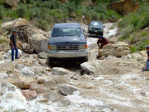 1987 Ford Bronco - A blue 1987 Ford Bronco tackles a challenging off-road path with another vehicle visible behind it.