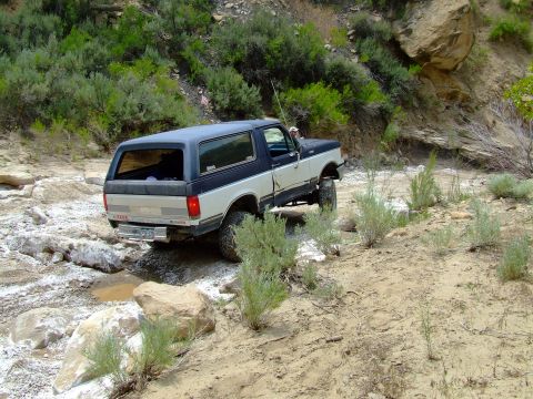 1987 Ford Bronco - A two-tone 1987 Ford Bronco navigates a challenging off-road course with rocky terrain and sparse vegetation.