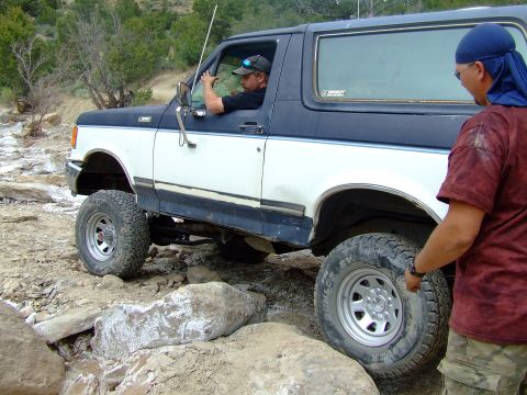 1987 Ford Bronco - A two-tone Ford Bronco is shown navigating a rocky trail, with one person driving and another observing.