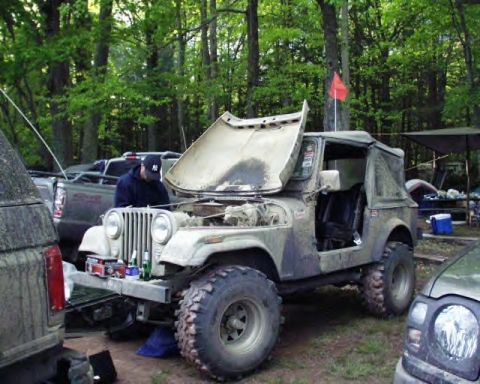 1991 Ford Bronco - A man works on a camouflaged Jeep CJ-7 with its hood open, parked in a wooded area.