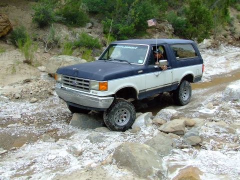 1987 Ford Bronco - A blue and white 1987 Ford Bronco navigates a rocky trail, displaying an American flag on its antenna.