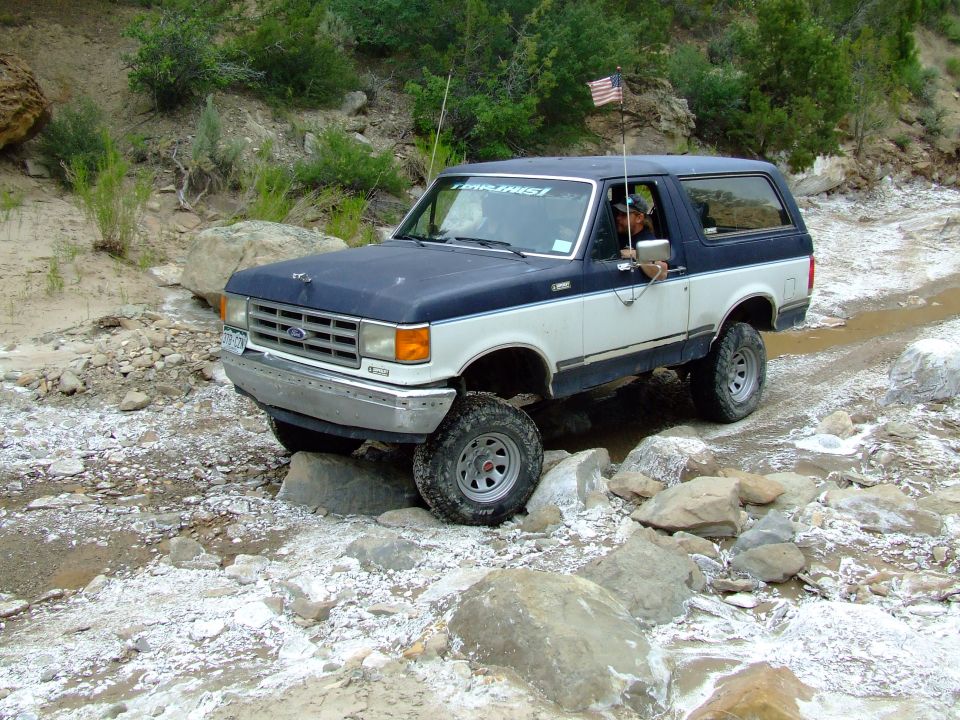 Blue and white 1987 Ford Bronco driving over rocks on a dirt path