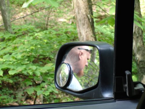1996 Ford Bronco - A reflection in the side mirror shows a man with a beard and sunglasses, set against a backdrop of green foliage and trees.