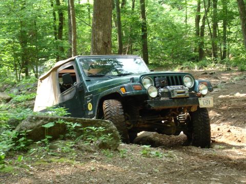 1996 Ford Bronco - A green Jeep Wrangler is shown off-roading through a wooded area with rocky terrain.